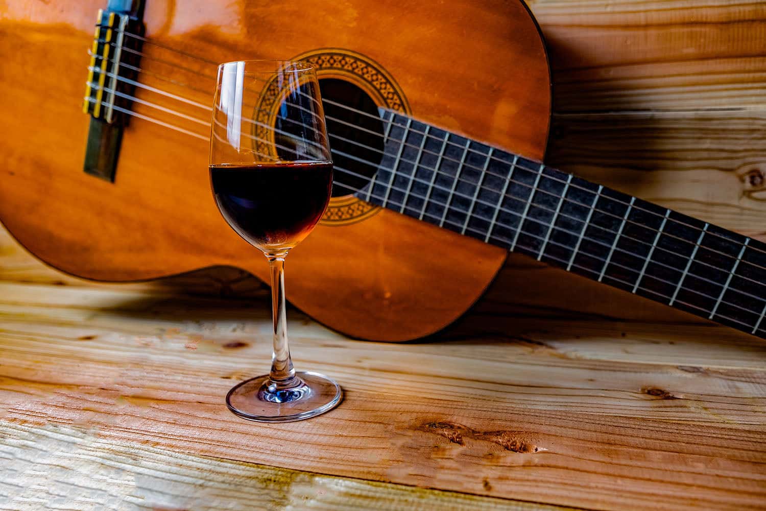 Glass of red wine on a wooden table in front of a classical guitar, evoking a rustic, musical ambiance.