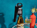 Wine bottle topped with a sleek black wine opener, next to a glass of white wine and a red vase with flowers.