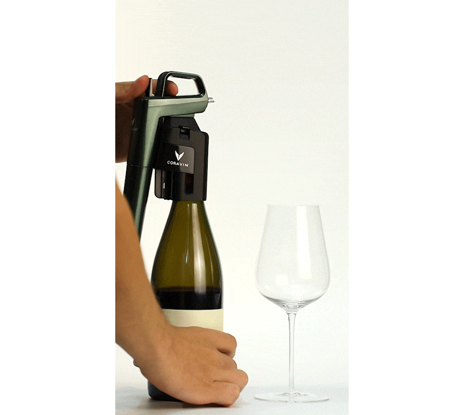 A hand uses a Coravin wine opener on a bottle beside an empty wine glass.