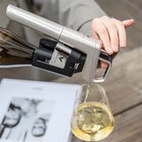 A hand pouring white wine into a glass using a sleek wine opener, with a magazine visible in the background.