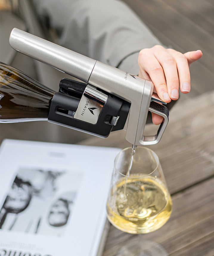 A hand pouring white wine into a glass using a sleek wine opener, with a magazine visible in the background.