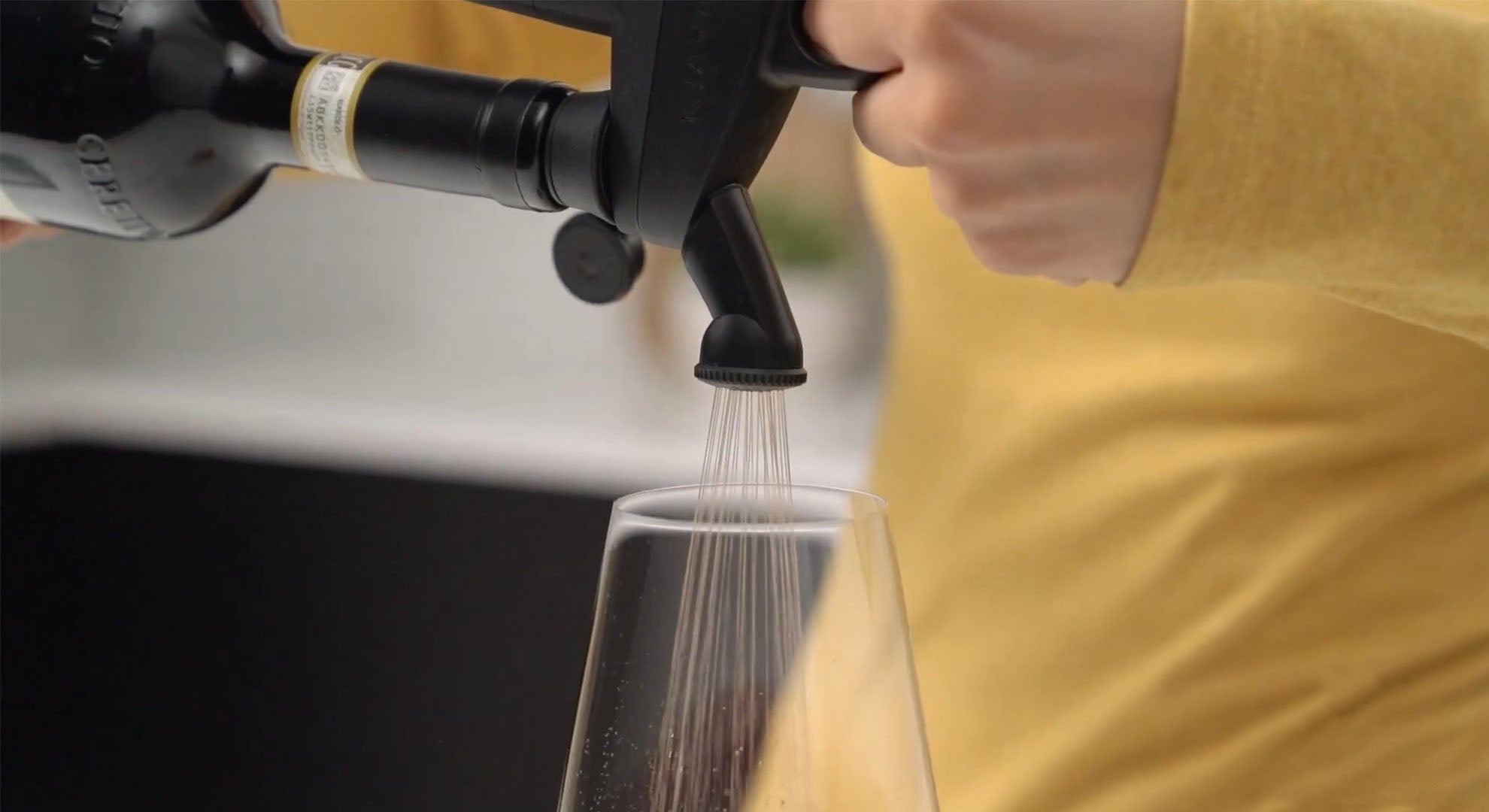 A hand pours wine from a bottle into a glass using a pouring tool, creating a fine spray effect.