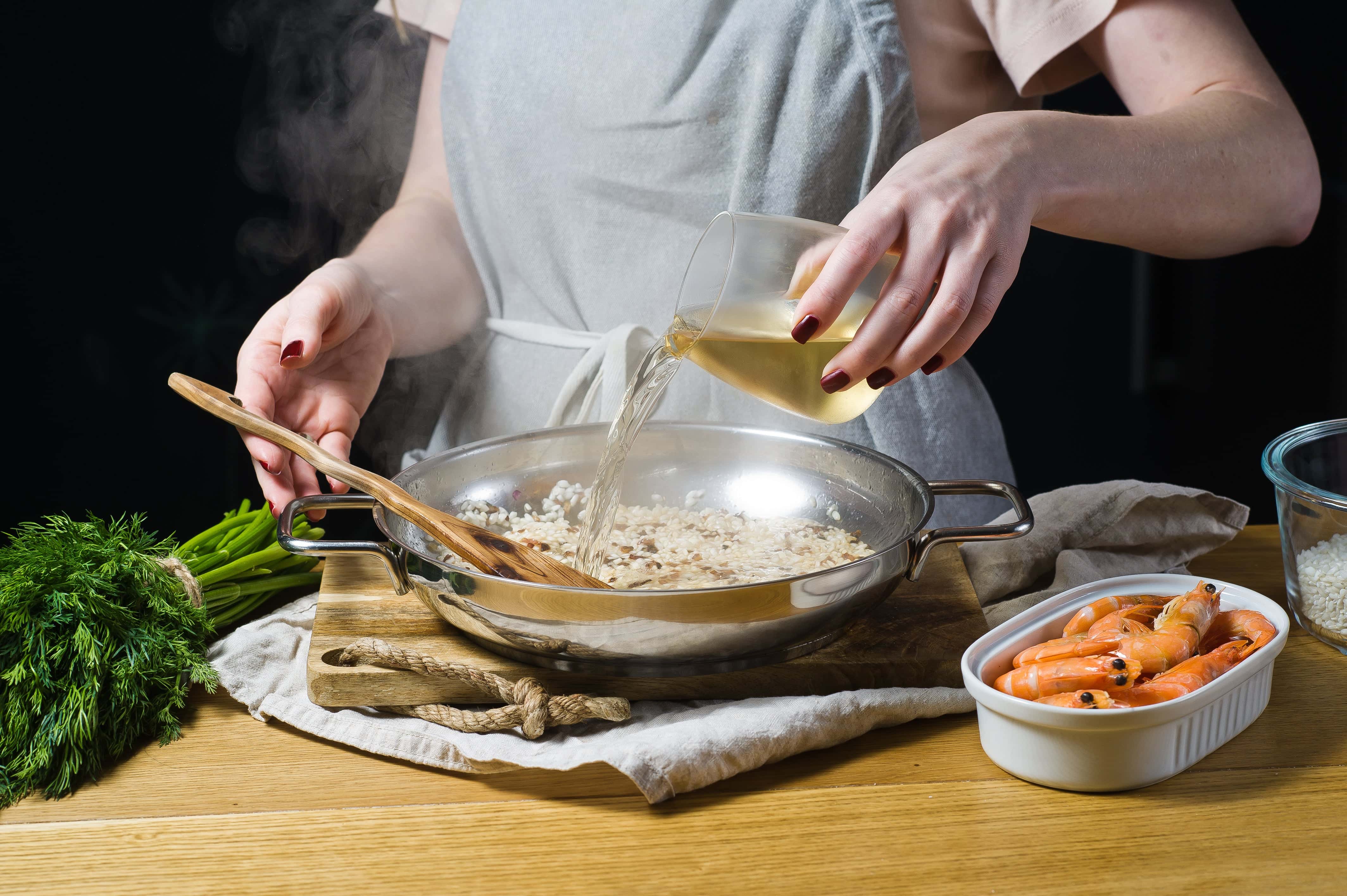 Woman in a grey apron pouring white wine into a steaming pan of risotto, with shrimp, herbs, and rice ingredients on the wooden table