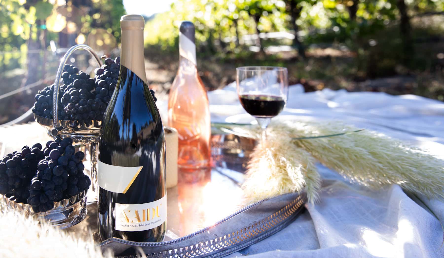 Naidu Wines Pinot Noir bottle with fresh grapes, rosé wine, and a glass of red wine set on a picnic tray in a sunlit vineyard.