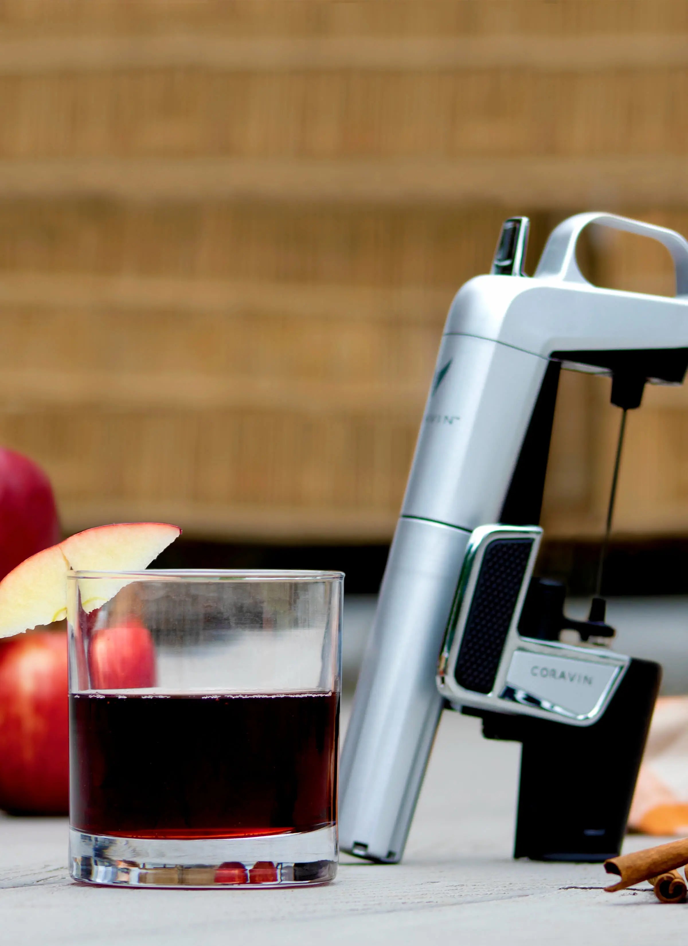 A glass of red wine garnished with an apple slice next to a Coravin wine preservation system, with apples and cinnamon sticks in the background on a wooden surface.
