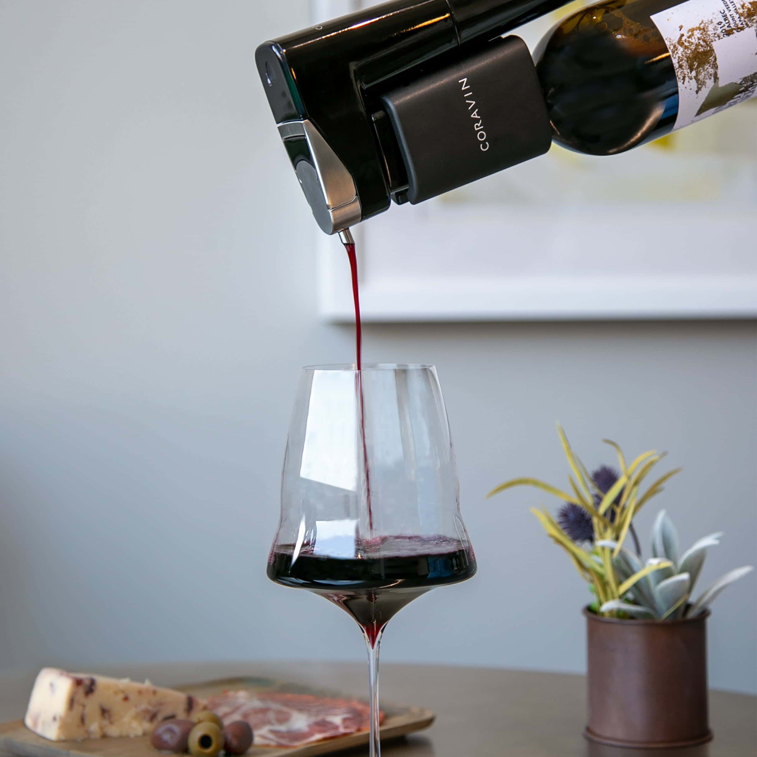 A Coravin wine preservation system pouring red wine into a modern stemmed glass, with a charcuterie board and potted plant in the background on a sleek tabletop.