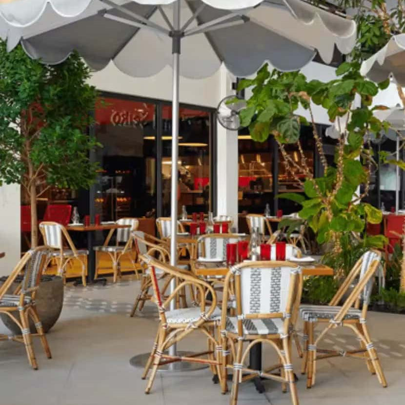 Outdoor café patio with wicker chairs and wooden tables under large white umbrellas, surrounded by lush green potted plants and adjacent to a modern restaurant entrance.