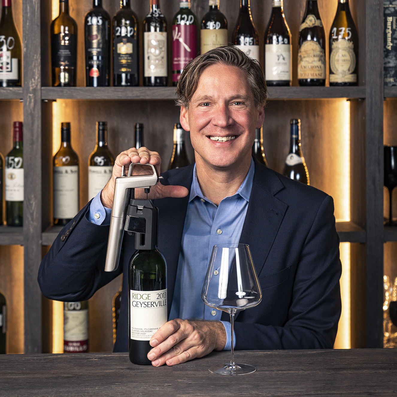 Smiling man in blazer demonstrating Coravin wine preservation system with Ridge Geyserville bottle in wine cellar.