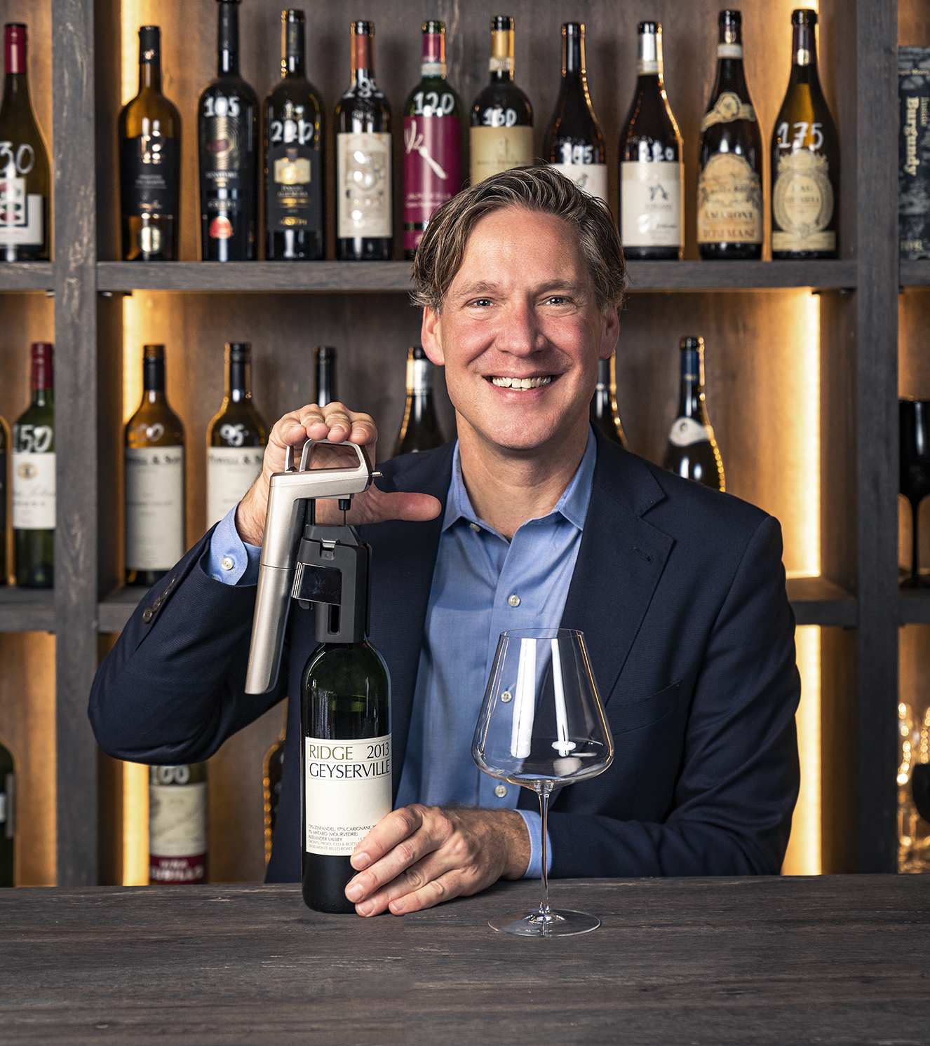 Smiling man in blazer demonstrating Coravin wine preservation system with Ridge Geyserville bottle in wine cellar.