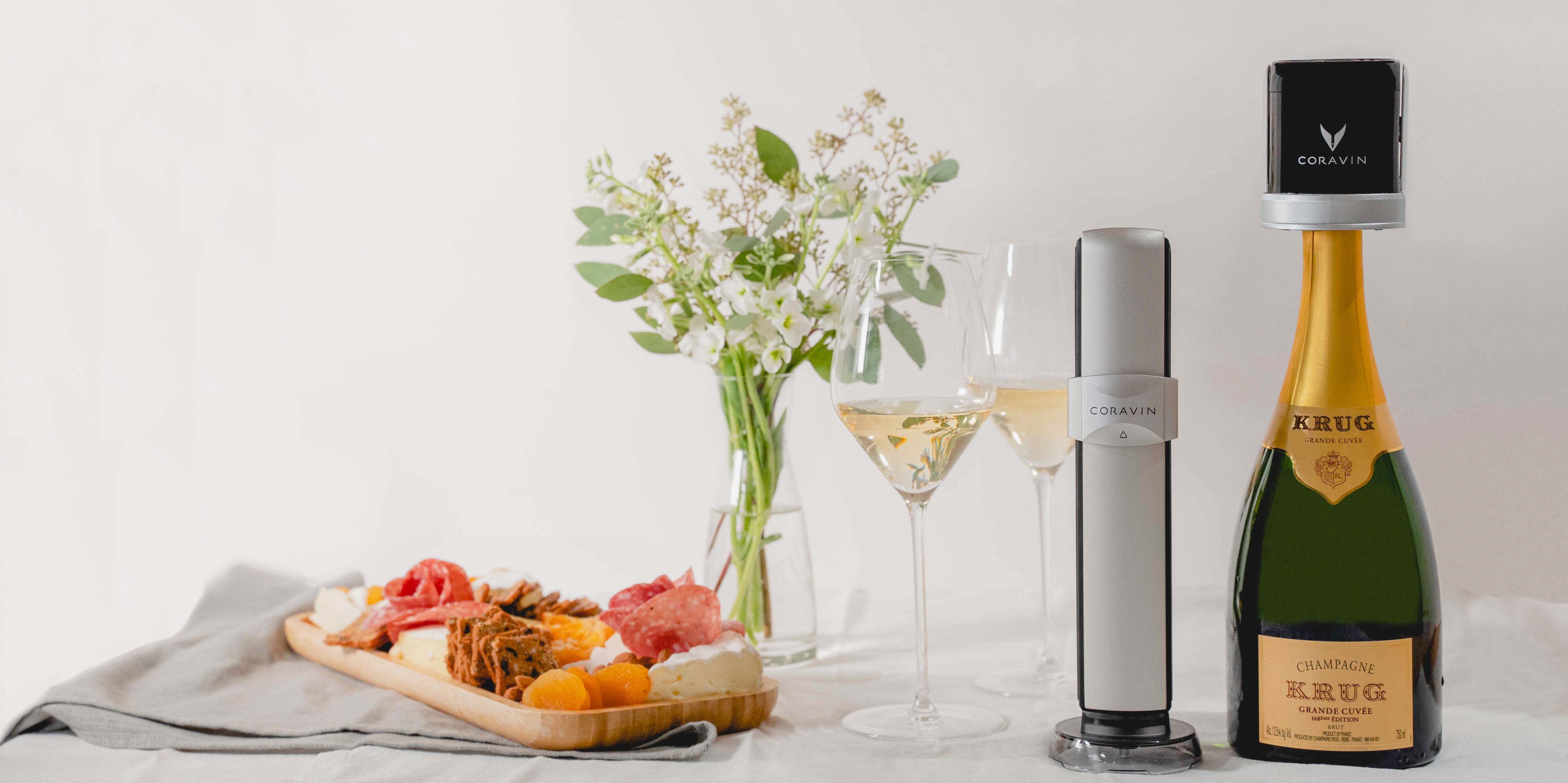 Coravin Sparkling Wine Preservation System with a bottle of Krug Grande Cuvée champagne, two glasses of sparkling wine, a charcuterie board, and a floral centerpiece on a white table.