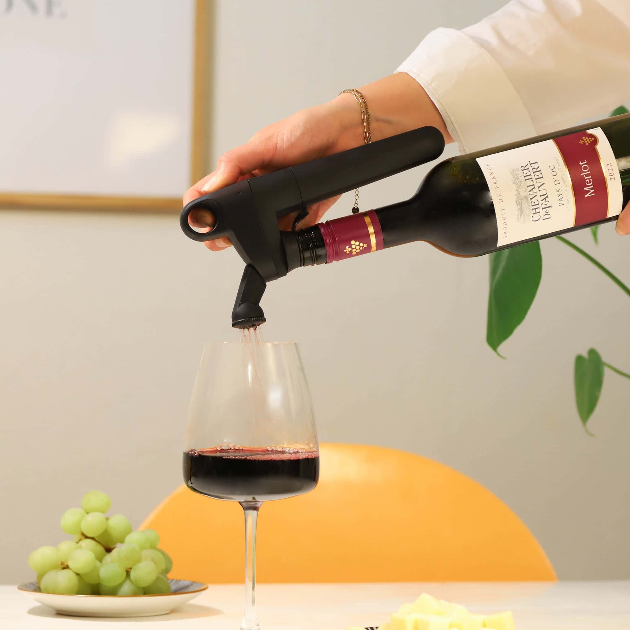 Pouring red wine from a Coravin Pivot Wine Preservation System into a glass, with grapes and cheese on a table, showcasing wine aeration and preservation.