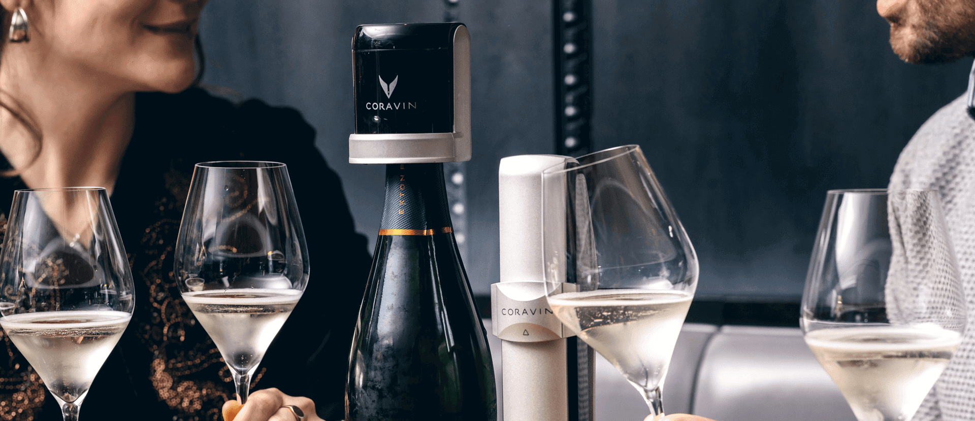 Two people enjoying glasses of sparkling wine with a Coravin Sparkling system preserving an open champagne bottle on the table between them.