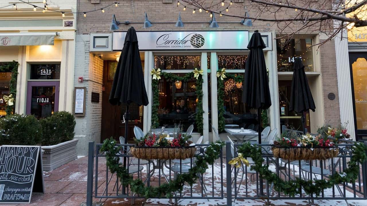 Corridor 44 Champagne, Cocktails, and Dining restaurant exterior decorated for the holidays with garlands, gold bows, and festive lights, featuring outdoor seating with black umbrellas and a 'Happy Hour' sign on the snow-dusted sidewalk.