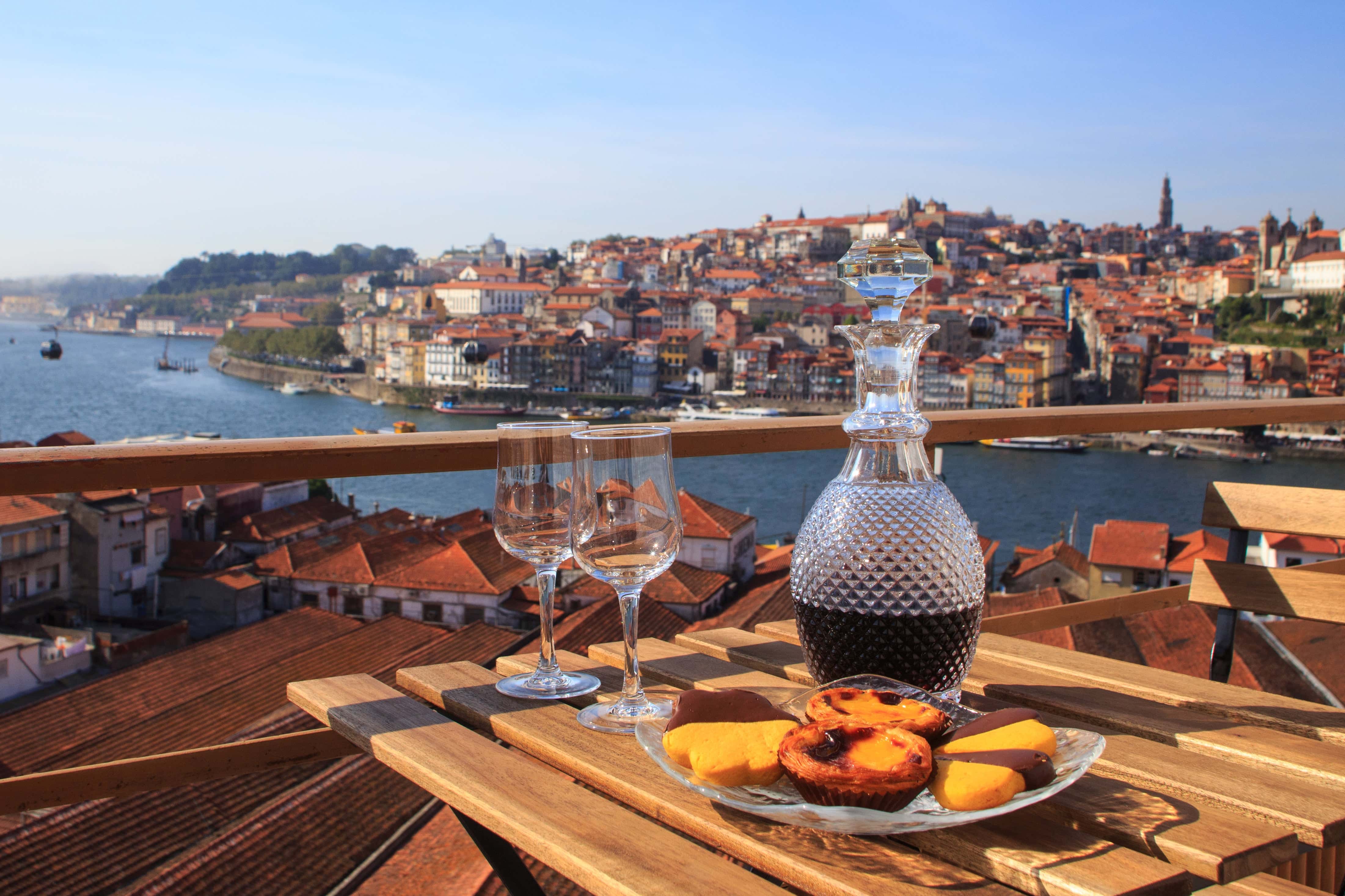 Decanter of port wine, two empty glasses, and traditional Portuguese pastries on a wooden table overlooking the Douro River and the historic cityscape of Porto, Portugal.