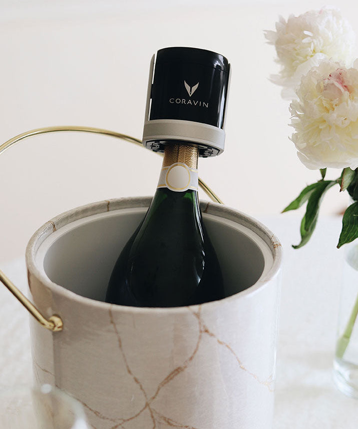 A bottle of champagne in a marble ice bucket, topped with a black Coravin wine preservation system. Peonies are visible nearby.